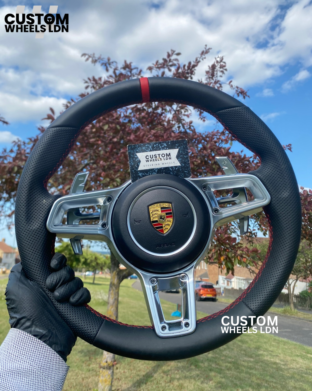 Steering wheel with Porsche logo held by a person outdoors, branded 'Custom Wheels LDN'.
